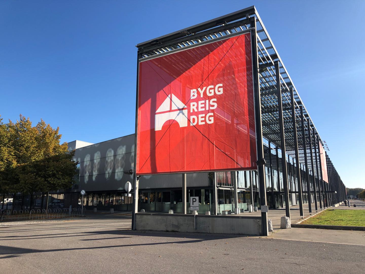 Fasade av et moderne kontorbygg med et stort, rektangulært banner som har teksten "BYGG REIS DEG" på en rød bakgrunn.