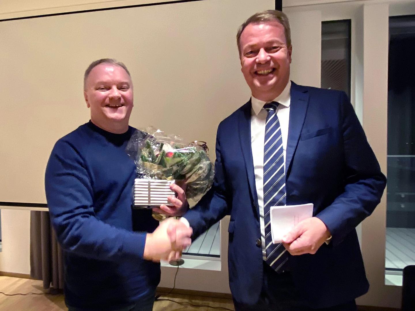 To personene håndhilser, én med blomster og gave, begge smilende i formell setting, kledd i forretningsantrekk.