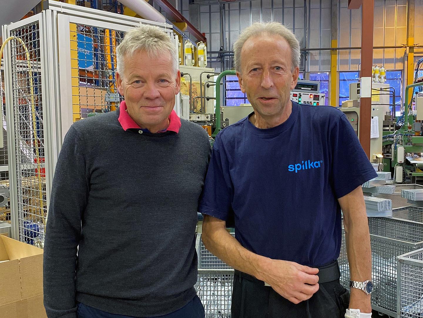 To menn står side om side i en fabrikk, omgitt av maskiner og utstyr, ser direkte mot kamera.