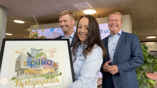 Tre personer smiler og holder et sertifikat under en signeringseremonie, mens de står i et innendørs miljø med planter og en skjerm i bakgrunnen.