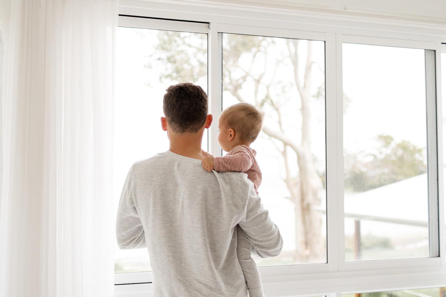 A father holding a child, looking out a white big window.