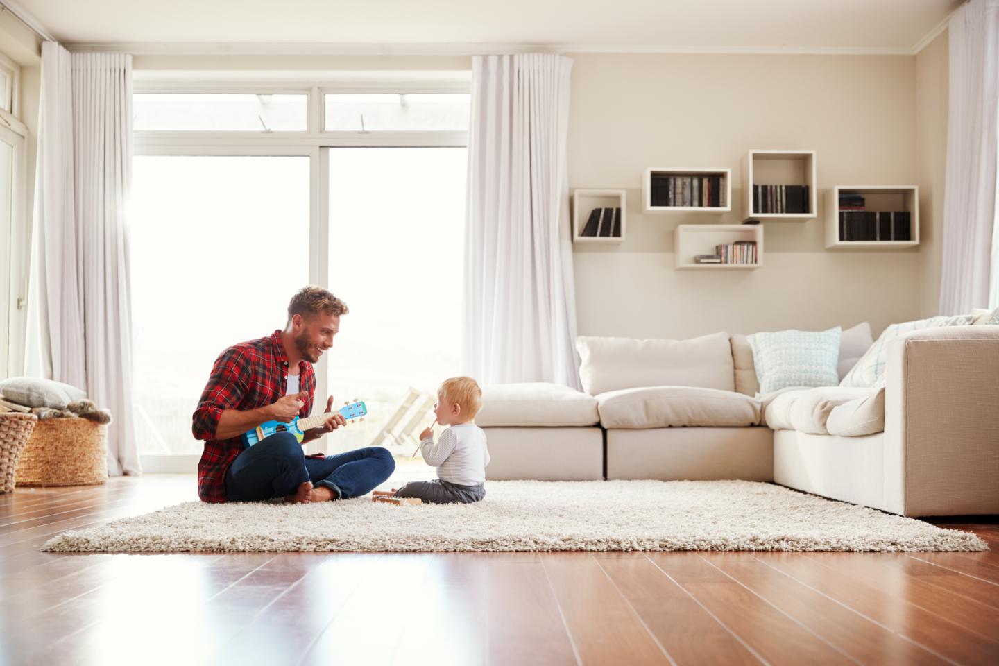 A man and a young child sit on a beige rug in a bright living room, playing with a small ukulele and engaging in a cheerful interaction.